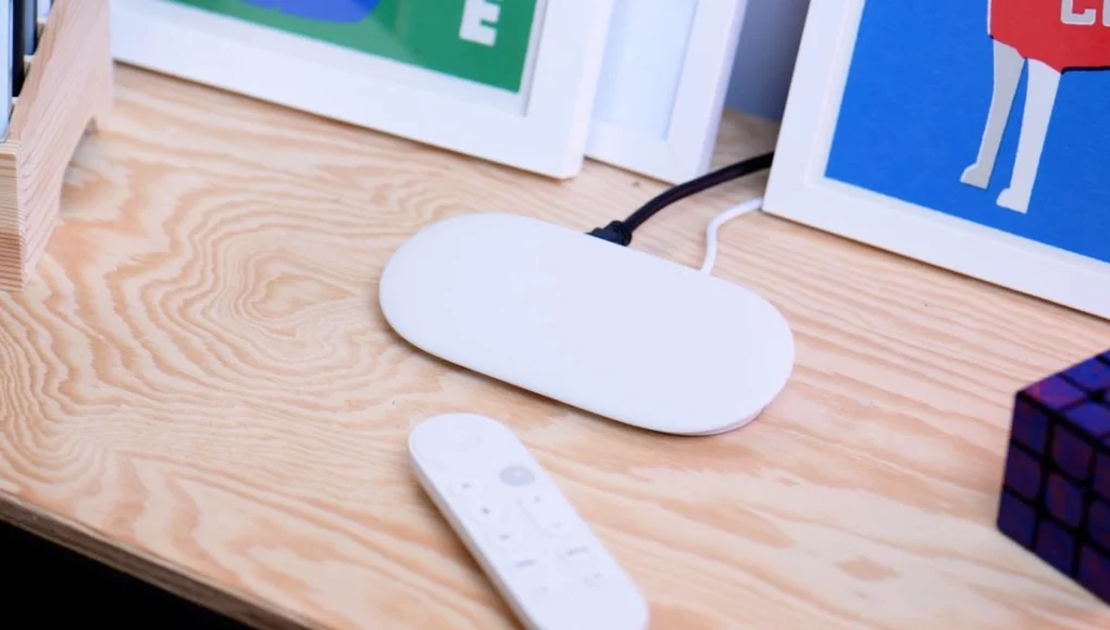 Dispositivo Google TV en color blanco sobre una mesa de madera, acompañado de un mando a distancia y un cubo tipo rompecabezas.
