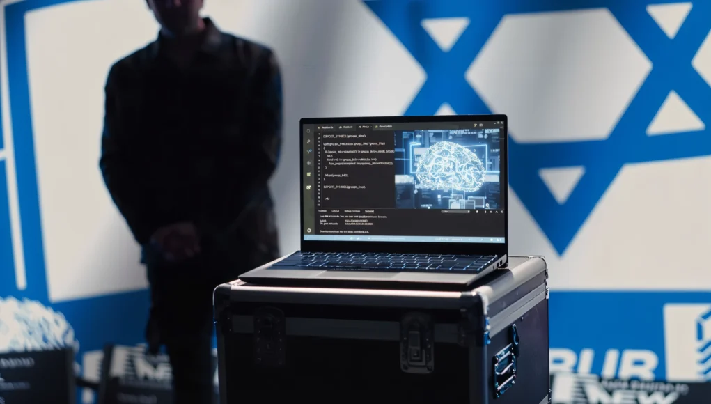Portátil abierto sobre una caja metálica mostrando en la pantalla un editor de código con líneas en C y, al lado, una ilustración digital de un cerebro con conexiones neuronales luminosas, representando inteligencia artificial. Al fondo se distingue de manera borrosa una persona y parte de la bandera de Israel en la pared.
