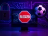 Bloqueos de LaLiga: Incongruencias políticas, abusos y vulneración de derechos fundamentales en España Ilustración digital en tonos azul y violeta que representa el bloqueo de internet. En el centro aparece un monitor con un cartel rojo de “BLOCKED” en forma de señal de stop, asegurado con una cadena. A la izquierda se ve un candado metálico y a la derecha una lupa enfocando un balón de fútbol, simbolizando la relación entre el fútbol y el bloqueo de servicios legítimos en la red.