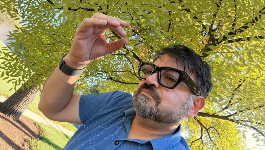 Hombre de barba y gafas grandes de color negro aparece en primer plano, levantando la mano con una pulsera electrónica en la muñeca. Está al aire libre, con un fondo de hojas verdes iluminadas por la luz del sol.