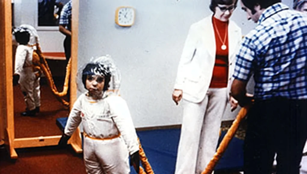 Niña con un traje protector especial conectado por un tubo a un sistema externo, utilizada en pruebas médicas durante la década de 1970, observada por dos adultos en una sala.