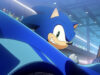 Sonic Racing: CrossWorlds no logra competir con Mario Kart World en las listas japonesas Ilustración animada de Sonic, el erizo azul, sonriendo con expresión confiada mientras conduce un vehículo de color azul en un entorno futurista iluminado con luces de neón y público al fondo.