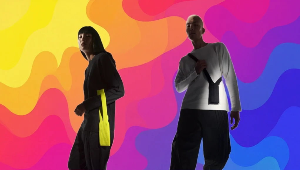 Dos personas posan con fundas textiles para iPhone en un fondo de colores degradados y ondulados que van del amarillo y naranja al rosa, púrpura y azul. La persona de la izquierda viste de negro con una funda amarilla colgando del brazo, y la de la derecha lleva una camiseta blanca con una funda negra cruzada al pecho.