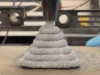 Investigadores desarrollan alternativa al cemento imprimible en 3D que endurece en tres días Una impresora 3D extruyendo capas gruesas de un material gris similar al cemento, formando una estructura cónica escalonada mientras un operario con guantes azules trabaja cerca del proceso.