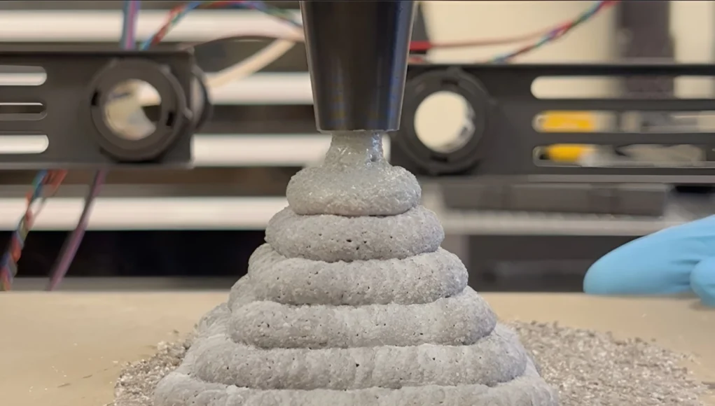 Una impresora 3D extruyendo capas gruesas de un material gris similar al cemento, formando una estructura cónica escalonada mientras un operario con guantes azules trabaja cerca del proceso.