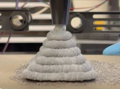 Investigadores desarrollan alternativa al cemento imprimible en 3D que endurece en tres días Una impresora 3D extruyendo capas gruesas de un material gris similar al cemento, formando una estructura cónica escalonada mientras un operario con guantes azules trabaja cerca del proceso.