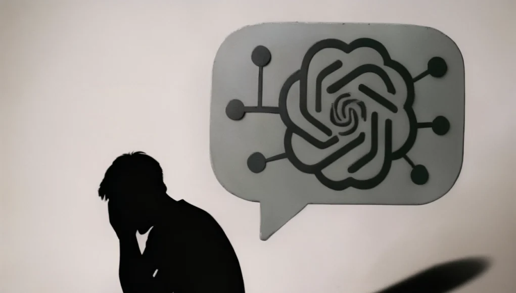 Silueta oscura de una persona sentada en el suelo, encorvada y con la cabeza entre las manos en señal de angustia. Frente a ella aparece un gran bocadillo de conversación gris con el logotipo estilizado de una inteligencia artificial y pequeñas conexiones a su alrededor, proyectando una sombra alargada. La escena es minimalista y transmite soledad, dependencia emocional y reflexión sobre la inteligencia artificial.