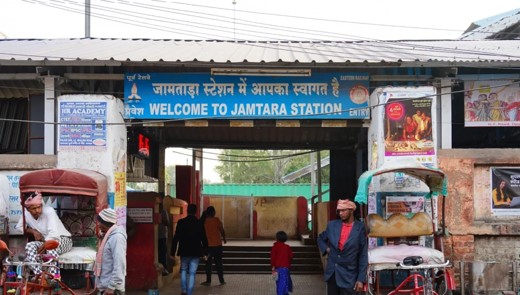 Entrada a la estación de tren de Jamtara, con un cartel que dice “Welcome to Jamtara Station” en inglés e hindi. Frente al acceso hay varias personas, rickshaws estacionados y vendedores locales.
