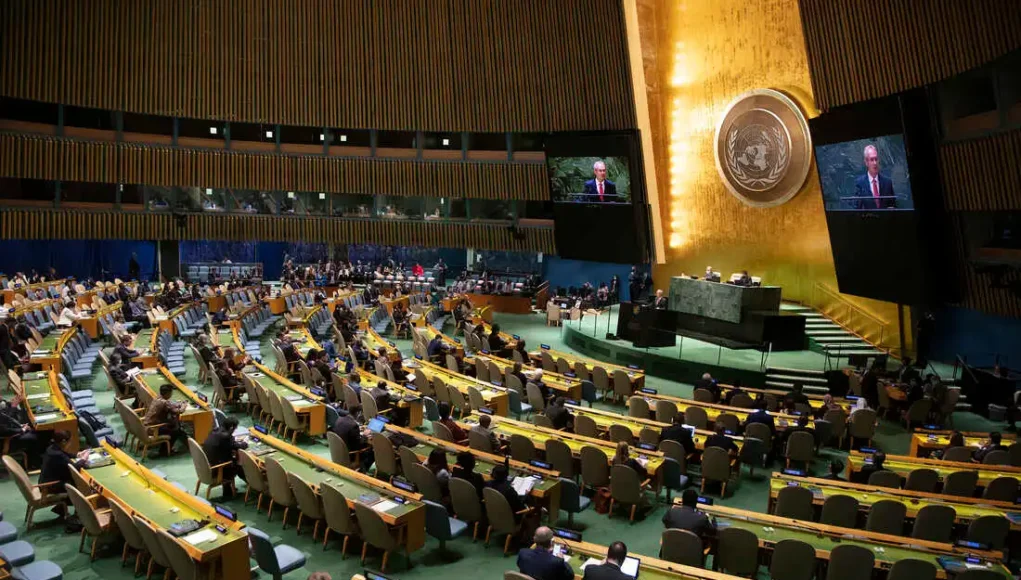 ONU Cibercrimen Vista general del salón de la Asamblea General de las Naciones Unidas, con delegados sentados en filas curvadas mientras escuchan un discurso desde el podio central. En el fondo se observa el emblema dorado de la ONU y grandes pantallas que muestran al orador.