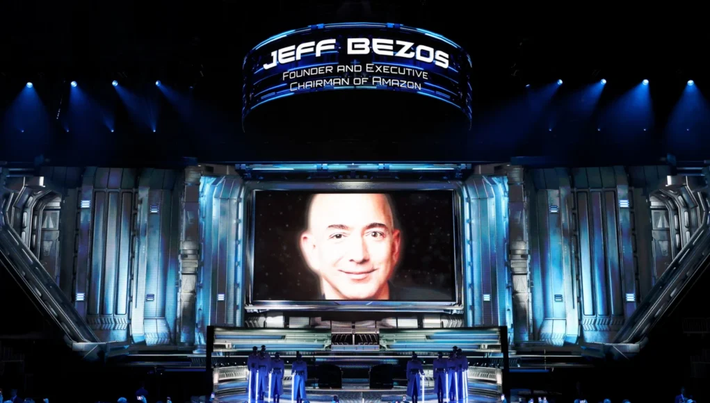 Un gran escenario futurista iluminado en tonos azules, con un público numeroso tomando fotos. En el centro hay una enorme pantalla que muestra el rostro de un hombre calvo sonriendo ligeramente. Sobre la pantalla aparece un cartel luminoso con texto que identifica a esa persona y su cargo. En la parte inferior, varias figuras uniformadas están alineadas en formación sobre la plataforma principal.