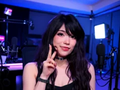Twitch donará $100,000 a organización benéfica tras el asalto a streamer Emiru en TwitchCon Una mujer joven posa en un estudio de grabación iluminado con tonos azulados y violetas. Lleva maquillaje marcado, un collar con forma de corazón y una pulsera con tachuelas. Sonríe a cámara mientras hace un gesto de paz con la mano; a su alrededor se ven micrófonos, brazos articulados y luces de estudio.