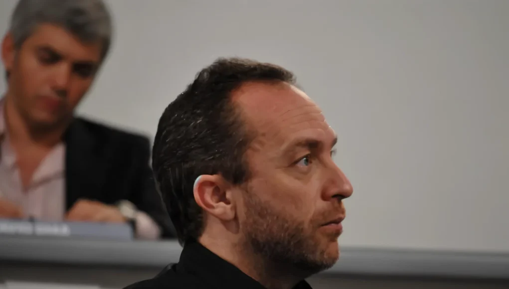 Hombre de mediana edad, con barba corta y cabello corto peinado hacia atrás, observado de perfil mientras asiste a un evento o conferencia. Viste ropa oscura y aparece concentrado mirando hacia el frente. En el fondo, otra persona está sentada, ligeramente desenfocada.