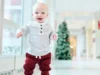 Bebé tratado con terapia génica CRISPR pionera da sus primeros pasos en casa por Navidad Bebé sonriente dando sus primeros pasos en un pasillo luminoso, vestido con una camiseta de rayas claras y pantalones rojos, en un entorno interior moderno y desenfocado.