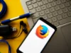 Mozilla confirma que Firefox evolucionará hacia un navegador con IA y los usuarios se enfadan Teléfono móvil mostrando el logotipo de Firefox sobre una pantalla blanca, apoyado junto a un ordenador portátil y varios accesorios tecnológicos como auriculares, cables USB y un bolígrafo, sobre un fondo amarillo que transmite un entorno de trabajo digital y tecnológico.