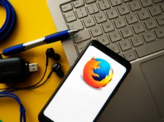 Mozilla confirma que Firefox evolucionará hacia un navegador con IA y los usuarios se enfadan Teléfono móvil mostrando el logotipo de Firefox sobre una pantalla blanca, apoyado junto a un ordenador portátil y varios accesorios tecnológicos como auriculares, cables USB y un bolígrafo, sobre un fondo amarillo que transmite un entorno de trabajo digital y tecnológico.
