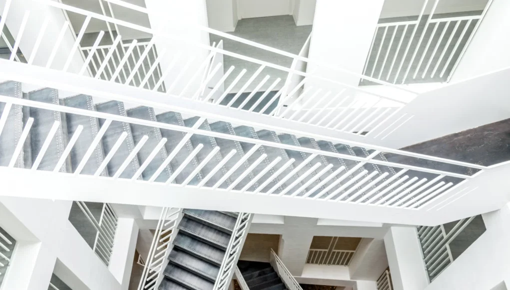 Vista cenital de un interior arquitectónico moderno con múltiples tramos de escaleras blancas y barandillas geométricas que se cruzan en distintos niveles, creando un patrón abstracto y simétrico de líneas y formas.