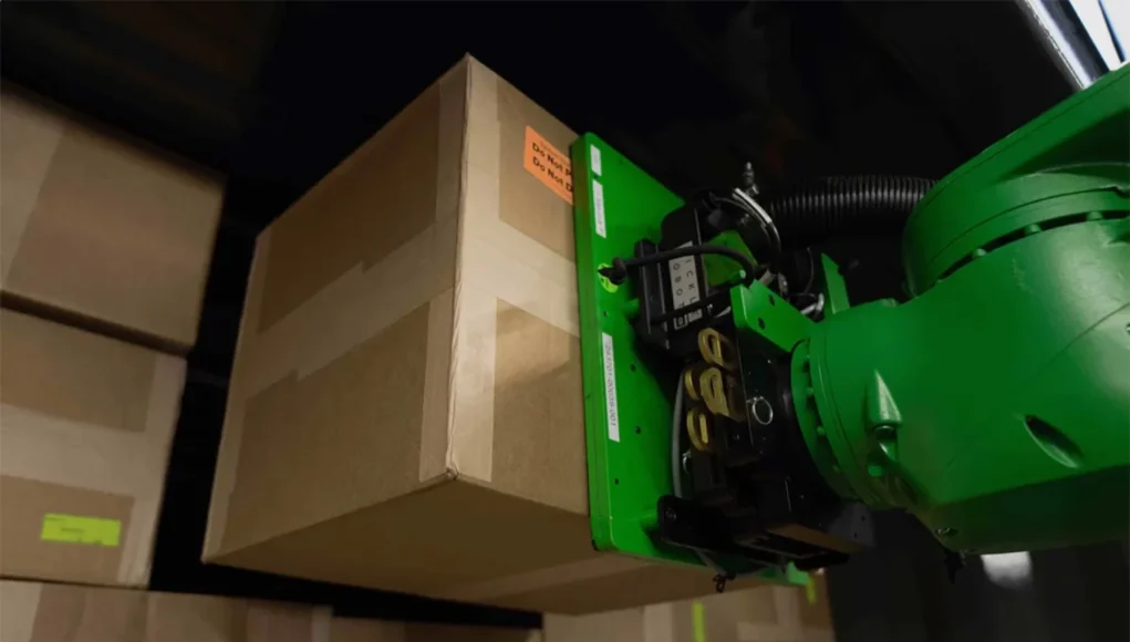 Un brazo robótico de color verde levanta una caja de cartón en un almacén lleno de paquetes apilados. El robot sostiene la caja mediante un mecanismo de agarre en su extremo.