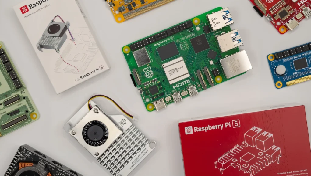 Varios componentes electrónicos dispuestos sobre una superficie clara, incluyendo una placa Raspberry Pi 5, cajas de embalaje, una unidad de refrigeración con ventilador y otras placas de expansión y accesorios.