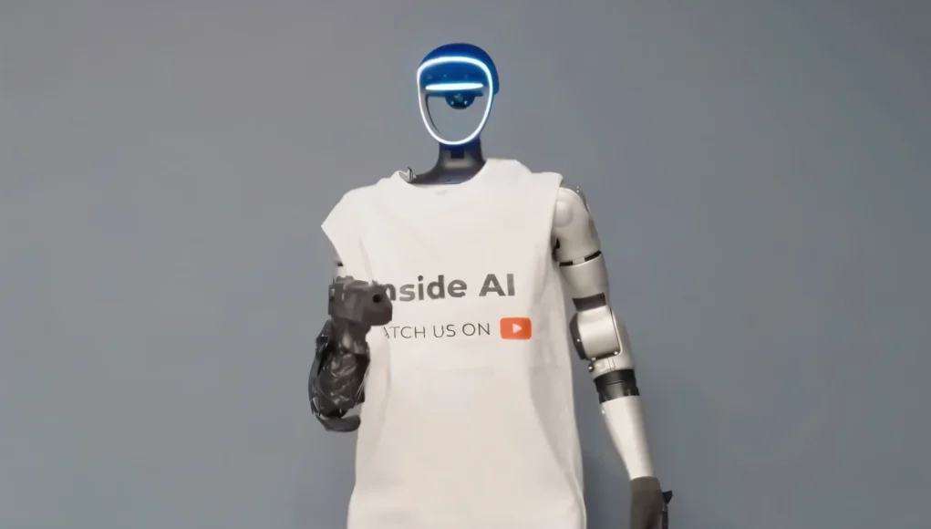 Robot humanoide de aspecto futurista con rostro iluminado en azul, vestido con una camiseta blanca con el texto “Inside AI” y un icono de YouTube, de pie frente a un fondo neutro gris, representando la divulgación y presencia de la inteligencia artificial en medios digitales.
