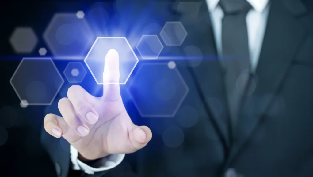 Mano de una persona con traje tocando una interfaz digital futurista, con un dedo presionando un icono hexagonal luminoso en tonos azules que representa autenticación o acceso tecnológico.
