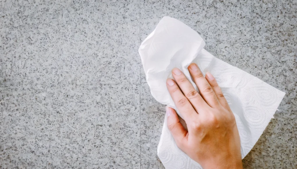 Mano limpiando una superficie de piedra o encimera gris con una servilleta de papel blanca, representando una acción de limpieza doméstica.
